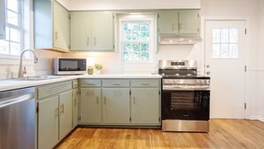 A kitchen with light green cabinets, a stainless steel oven, a microwave, and a window with a view of trees.