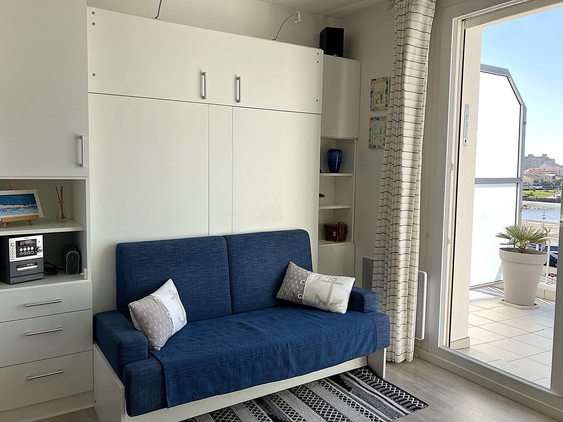 Una habitación con un sofá azul, armarios blancos, una mesita con un cuadro y vistas al exterior.