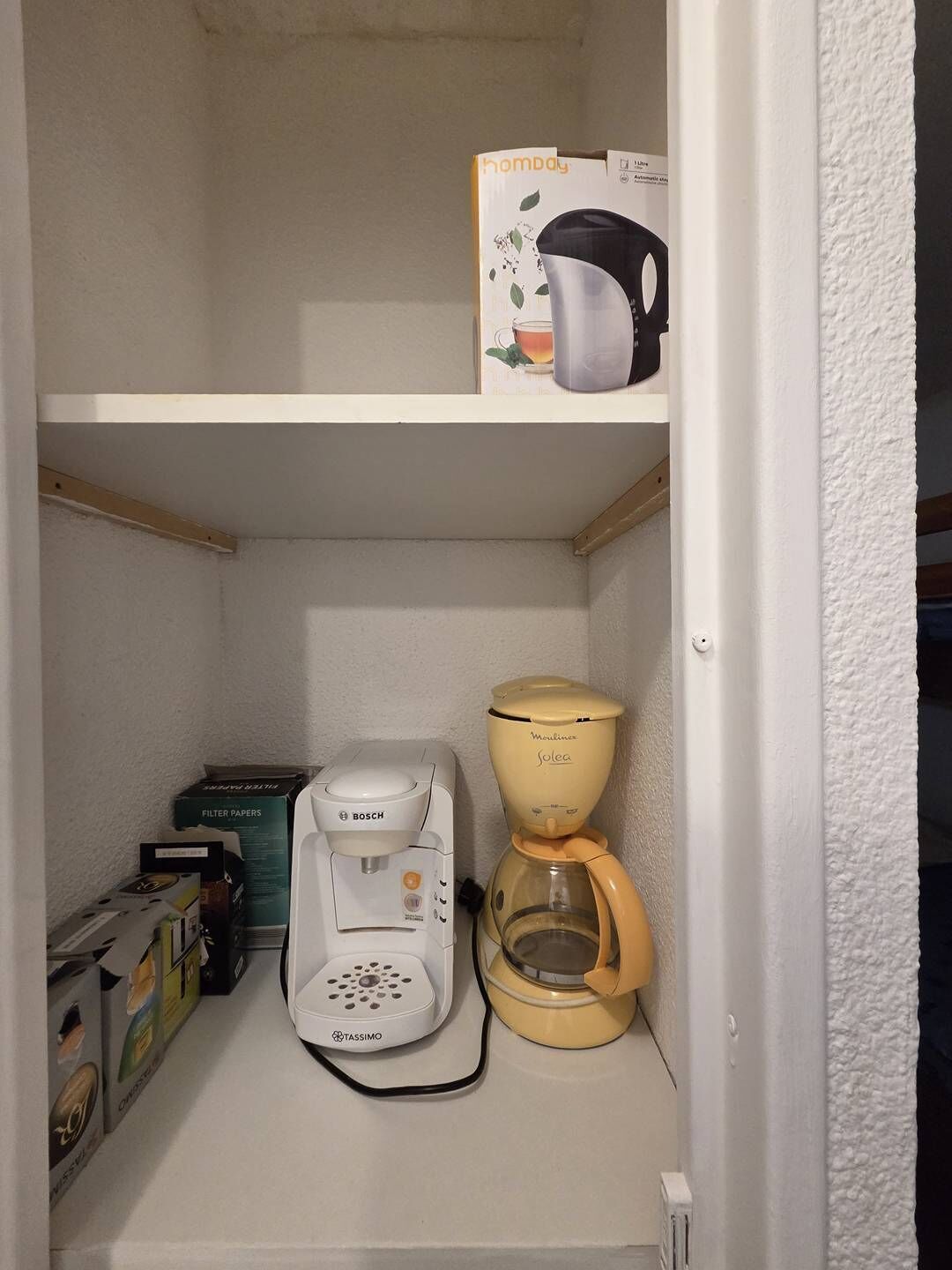 Una kitchenette pequeña con cafetera, jarra para café y cajas de filtro de papel.