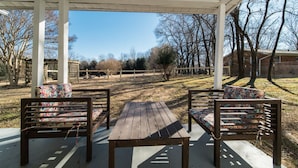 An outdoor patio with a wooden table and two chairs, surrounded by a white fence and trees.
