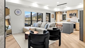 A modern living room with a sofa, two chairs, a coffee table, and a kitchen area with a refrigerator, microwave, and island. - 3BR Rockies Haven. Sleeps 8 with Hot Tub, Pool and Mountain Views (Canmore)