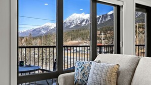 A balcony with a view of snow-capped mountains, a sofa with patterned cushions, and a small table with a blue tablecloth. - 3BR Rockies Haven. Sleeps 8 with Hot Tub, Pool and Mountain Views (Canmore)