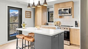 A modern kitchen with a central island, bar stools, a microwave, stove, and a view through a sliding door. - 3BR Rockies Haven. Sleeps 8 with Hot Tub, Pool and Mountain Views (Canmore)