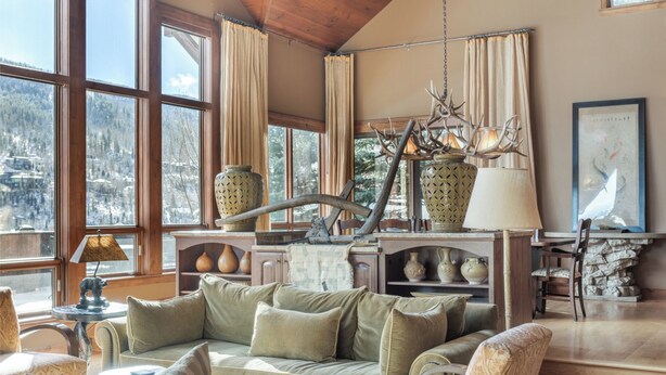 Un accogliente soggiorno con divano, poltrone, tavolino da caffè e camino. Grandi finestre con tende, un lampadario e diversi oggetti decorativi sugli scaffali.