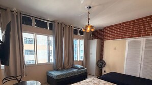 Ett kompakt hotellrum med en säng, ett litet bord med stolar, ett fönster med gardiner, en takfläkt och en vägg av tegelsten.
