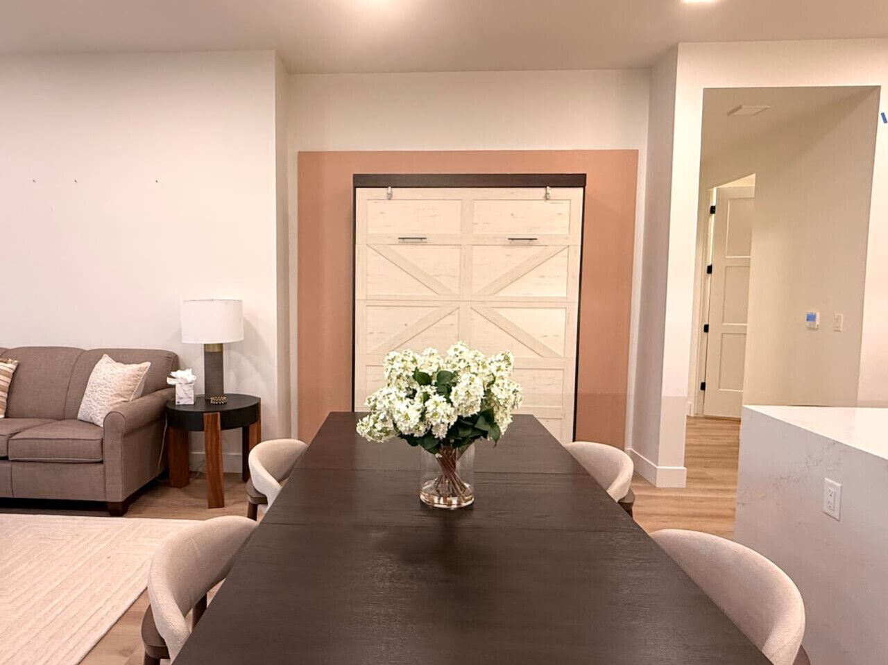A dining area with a dark wooden table, white chairs, a vase of white flowers, a lamp, and a sofa.