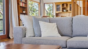 A grey sofa with three pillows, a wooden bookshelf, and a view of the outdoors through large windows. - Spacious Blue Mountains Retreat with Stunning Views (Mount Victoria)