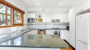 A modern kitchen with white cabinets, a granite countertop, stainless steel appliances, and a window with a view of trees. - Spacious Blue Mountains Retreat with Stunning Views (Mount Victoria)