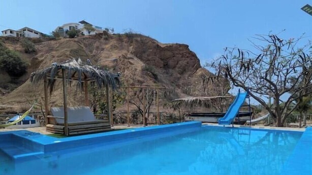 Une piscine avec un toboggan, un espace de détente ombragé et une vue sur une colline parsemée de bâtiments.