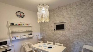 A dining area with a white table, chairs, and a chandelier. There is a white cabinet with shelves and a microwave on the left. A stone wall and a small TV are visible in the background. - Holiday flat with air conditioning and terrace (Eraclea Mare)