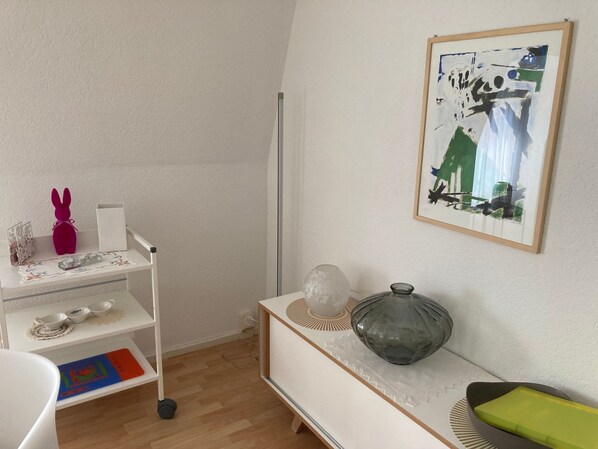 A room with a white wall, a framed abstract painting, a white shelf with decorative items, a white desk with a vase, and a colorful mat on the floor. - Domizil 1 by Interhome (Altstätten SG)