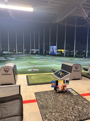 An indoor golf driving range with multiple practice stations, a control panel, and a seating area. - Miami Getaway - Stadium, Beaches & Nightlife (Miami)