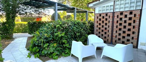 A patio with tiled flooring, white outdoor furniture, a brick wall, and a covered area with a metal frame.