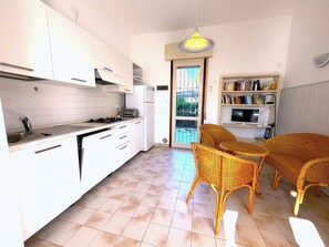 Cocina compacta con gabinetes blancos, estufa a gas y horno empotrado. Hay una zona de comedor con sillas de mimbre y una mesa pequeña. Se observa una estantería con libros y un televisor.