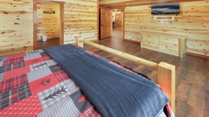 A log cabin interior with a bed, wooden walls, a ceiling fan, and a television on the wall. - Spacious 8-bedroom Cabin in the Smokies w/ Heated Pool, Hot Tub, & Movie/Game Rm (Sevierville)