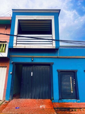 Un edificio de dos pisos con fachada azul, una puerta de garaje negra y una ventana pequeña.