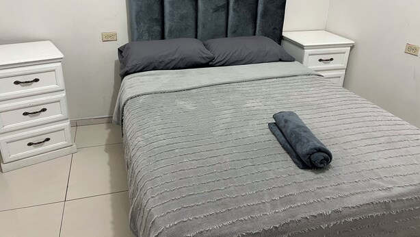 A neatly made bed with a grey bedspread and pillows, a grey upholstered headboard, and a white bedside table with drawers. - House 1 centrally located, 8 (up to 14 people) in san blas neighborhood, Comayagua (Comayagua)