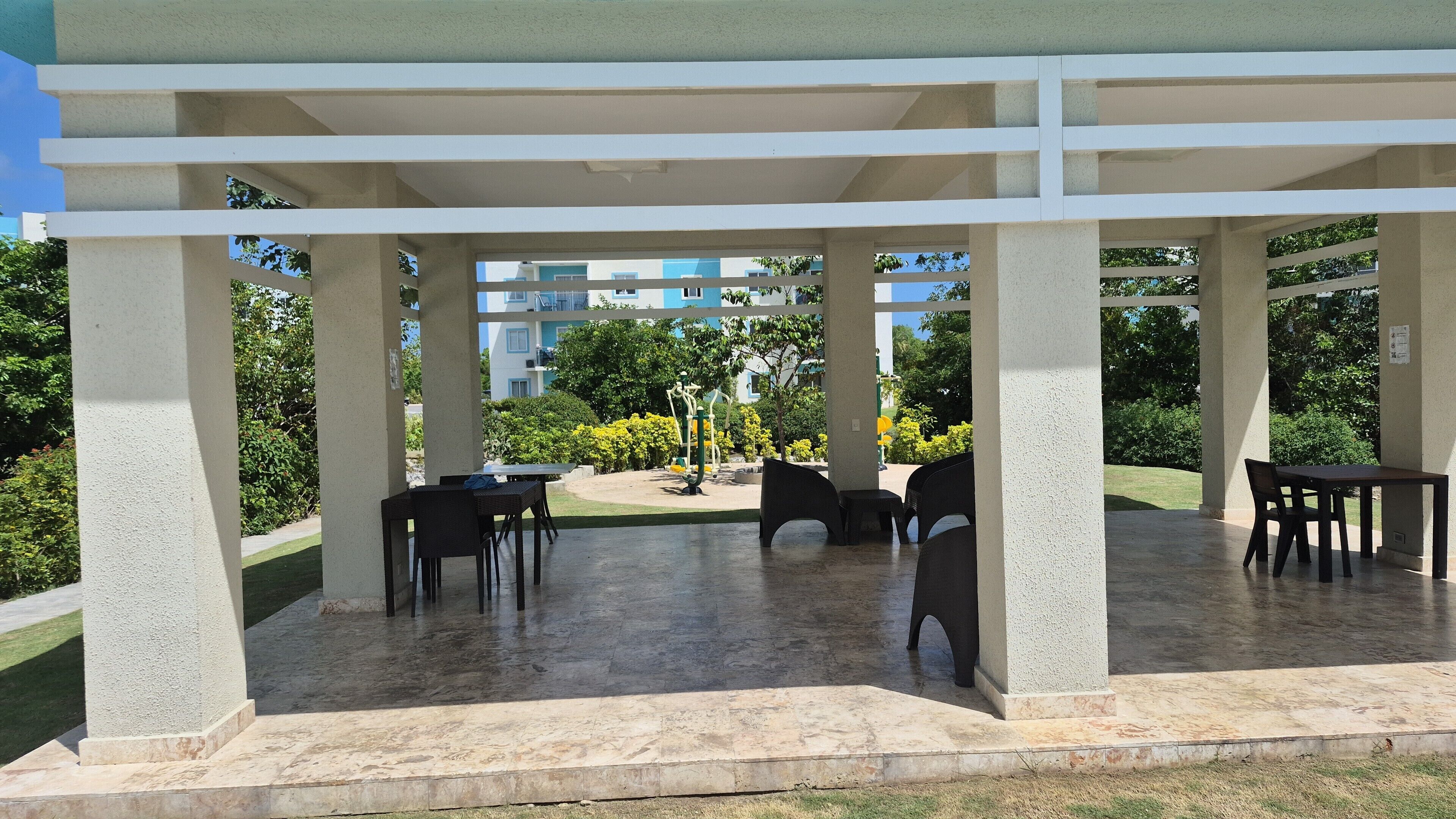 Un espace extérieur aménagé, abrité et doté de tables et de chaises, entouré de verdure et offrant une vue sur un bâtiment à la façade bleue et blanche.