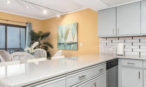 A modern kitchen with white cabinets, a marble countertop, and a painting of a sailboat. - Bayside Bliss | Sunset Views | Walk/Bike to Beach (Ocean City)