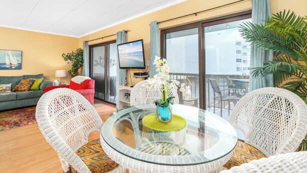 Un salon moderne avec une table en verre, des fauteuils en osier, un canapé rouge, un téléviseur et un balcon avec vue.