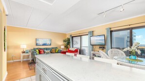 Une cuisine moderne dotée d’un grand îlot central, un coin repas avec une table en verre et un salon avec un canapé et un téléviseur.