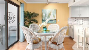 Un coin repas avec une table en verre entourée de chaises en osier, une plante en pot et un tableau représentant des voiliers accroché au mur.