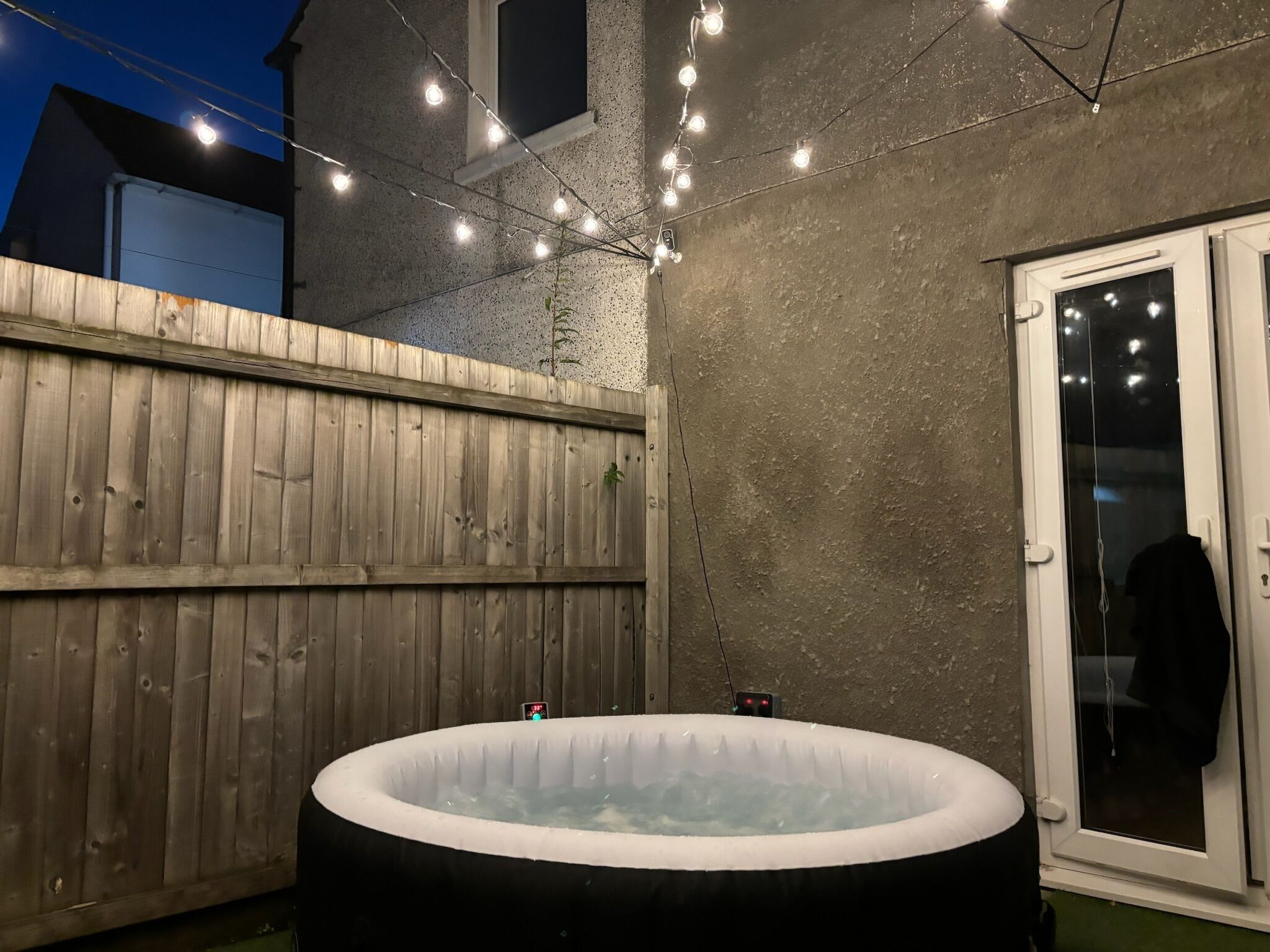 Ein Außenbereich mit Whirlpool, umzäunt durch einen Holzzaun, mit Lichterketten und einer Tür zu einem überdachten Bereich.