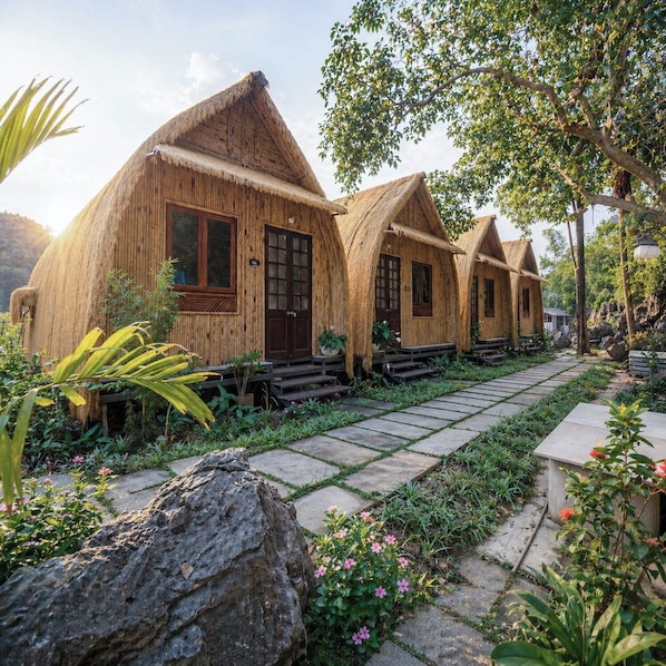 A row of thatched-roof cottages with a stone pathway leading to them, surrounded by greenery and rocks. - Tràng An Legend (Hoa Lu)