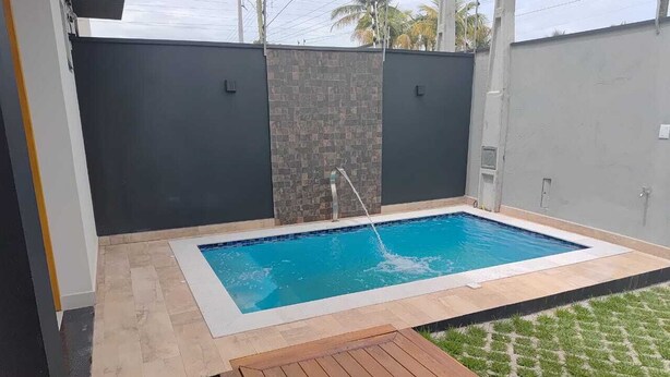 A rectangular swimming pool with a stone wall and a wooden deck in the foreground. - Refuge 1 Indaiá Beach Bertioga 150 meters from the sea (Bertioga)