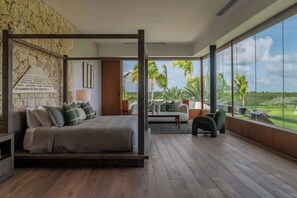Ein modernes Schlafzimmer mit Himmelbett, großem Fenster mit Blick auf Palmen und einer gemütlichen Sitzecke inklusive Sofa und Sesseln.