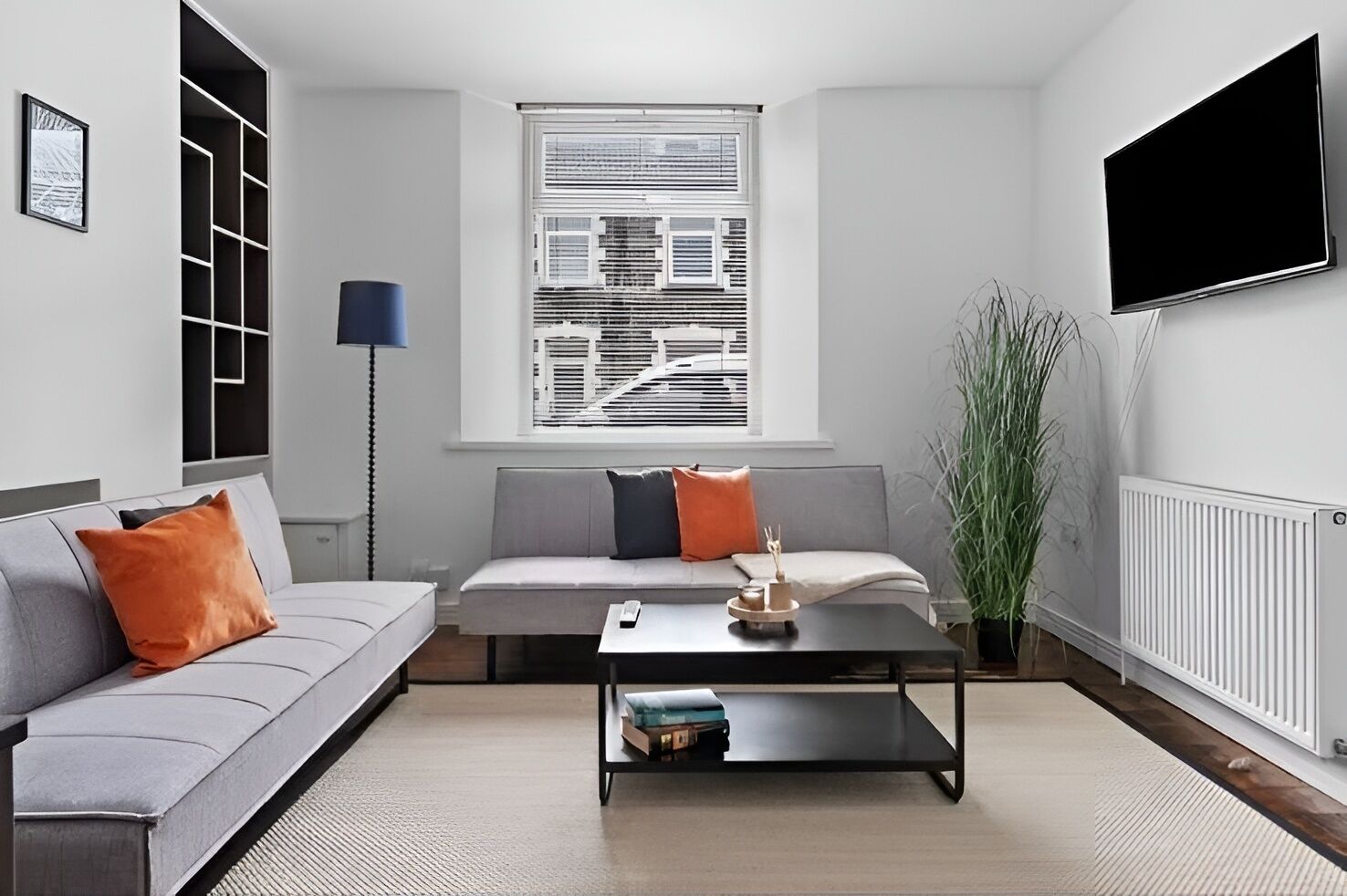 A modern living room with a grey sofa, a coffee table, a floor lamp, a large window with blinds, and a TV mounted on the wall.
