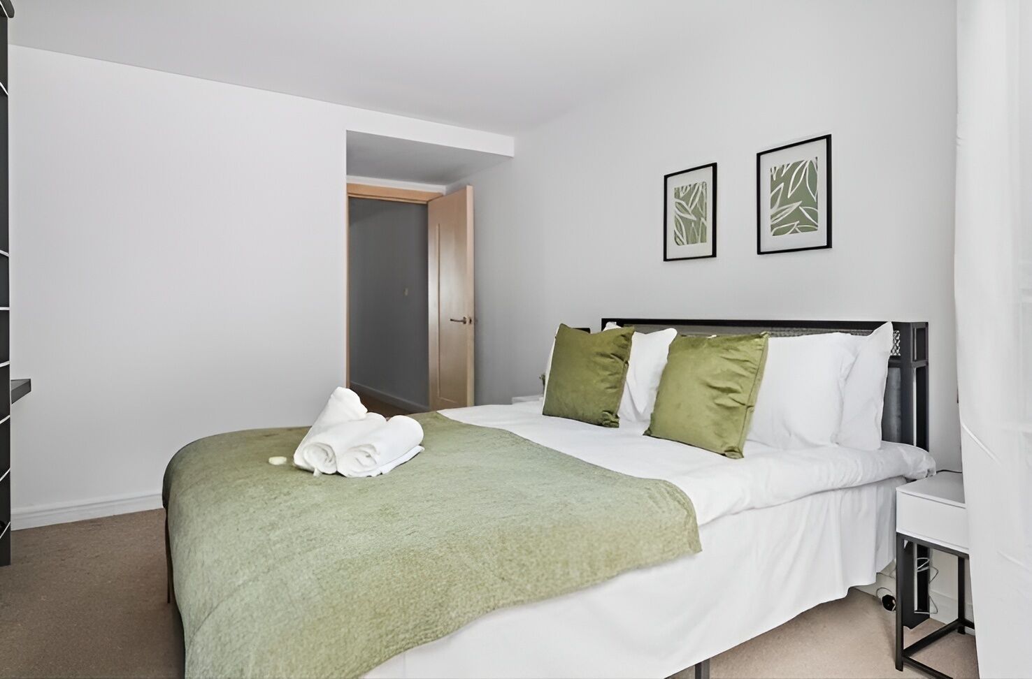 A neatly made bed with a green blanket and white pillows, a bedside table, and two framed pictures on the wall.