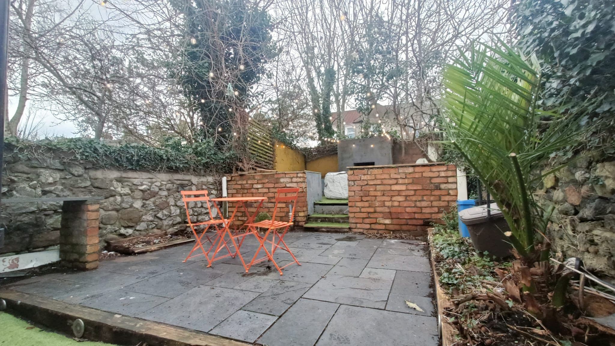 A patio area with a brick wall, a small table and chairs, and a potted plant.