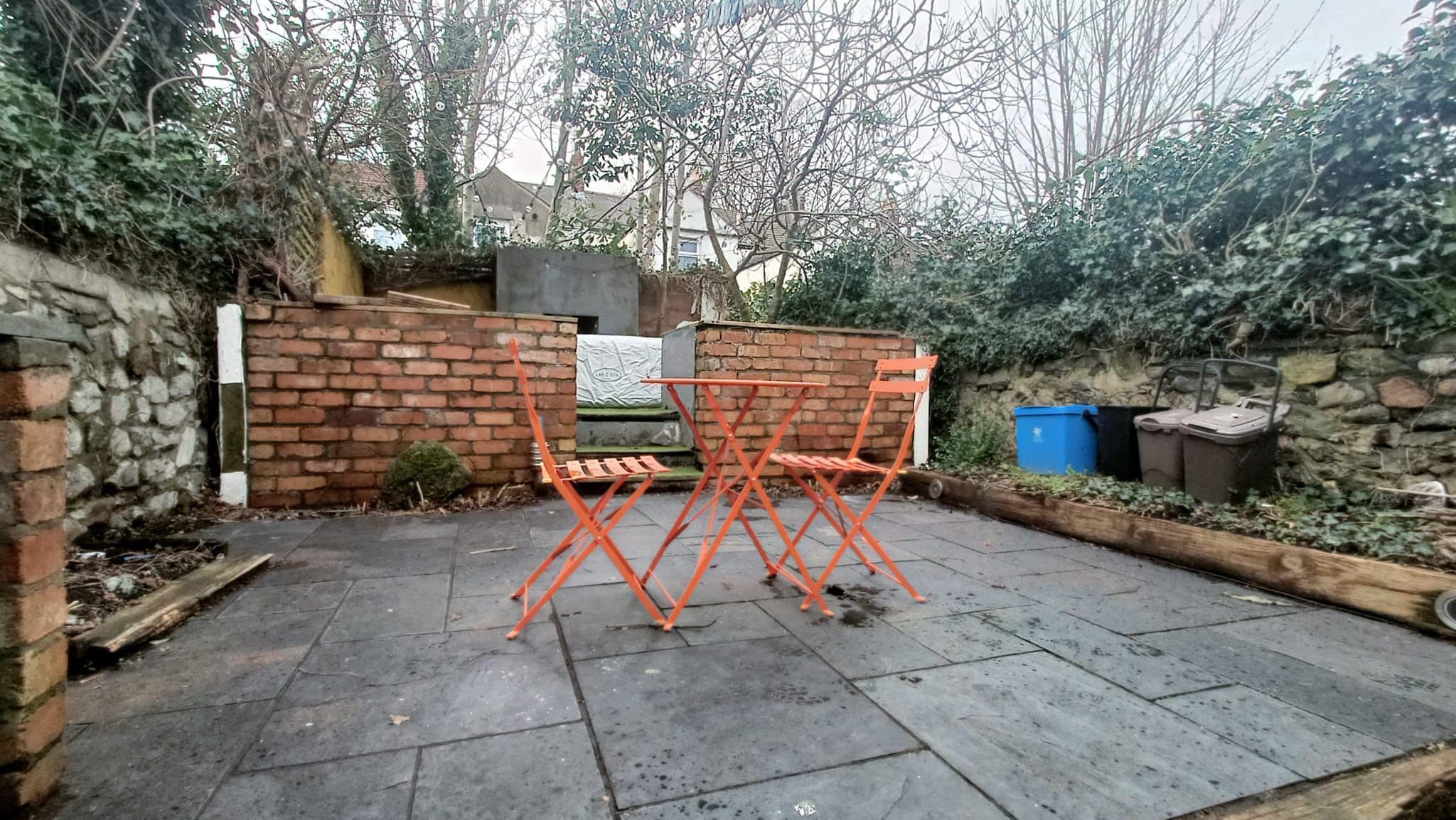 Une terrasse avec deux chaises pliantes orange, un mur de briques et un mur de pierres, ainsi qu’une poubelle bleue.