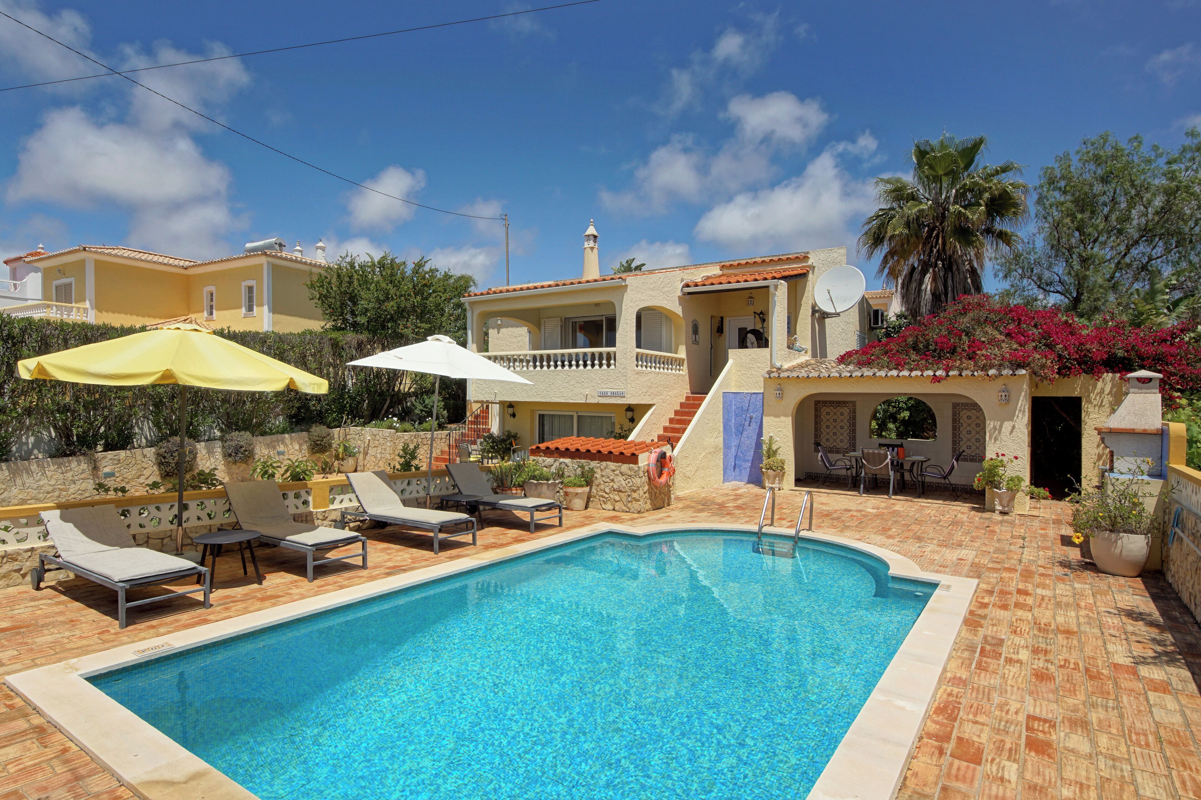 A swimming pool with lounge chairs and umbrellas, surrounded by a stone wall and a building with a tiled roof.
