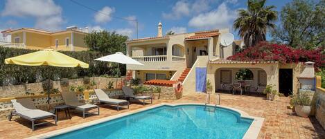 A swimming pool with lounge chairs and umbrellas, surrounded by a stone wall and a building with a tiled roof.