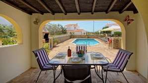 Un porche cubierto con una mesa para dos, con vista a una piscina y una villa.