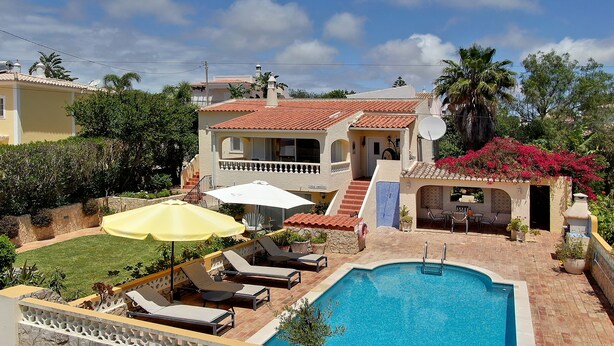 Zona de piscina con sillones de descanso y sombrillas, piscina y un pequeño patio con una parrilla.