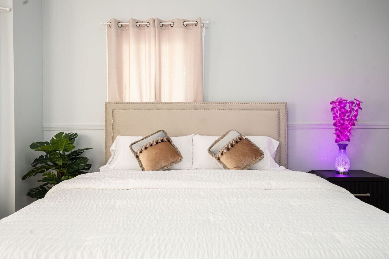 A neatly made bed with white bedding and two decorative pillows, a bedside plant, and a purple vase on a nightstand.