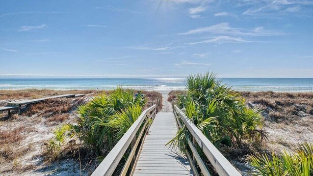 Ein hölzerner Steg führt zu einem Strand mit blauem Himmel und Blick auf das Meer.