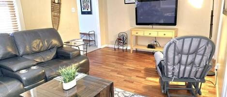 Un salon avec un canapé en cuir, une table basse en bois agrémentée d’une plante, un fauteuil gris, un téléviseur fixé au mur et une console jaune.
