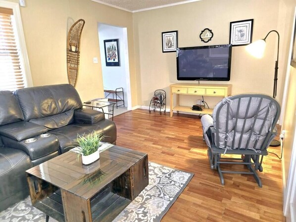 Ein Wohnzimmer mit einem Ledersofa, einem Couchtisch aus Holz mit einer Pflanze, einem grauen Sessel, einem Fernseher an der Wand und einem gelben Konsolentisch.