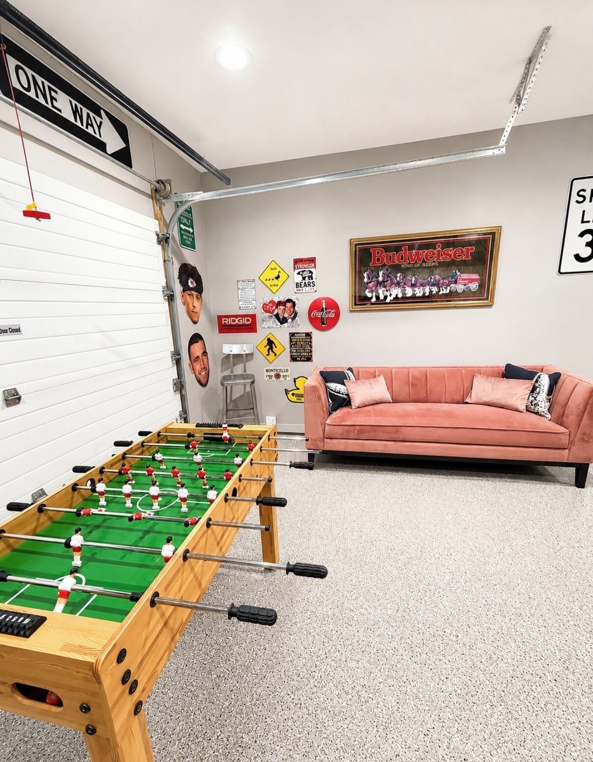 Ein Zimmer mit einem Fußballtisch, einem rosa Sofa und verschiedenen Wanddekorationen, darunter ein Budweiser-Schild und ein 'ONE WAY'-Schild.