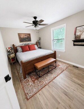 Quarto com cama, um banquinho, ventoinha no teto, janela com persianas e prateleira fixada na parede.