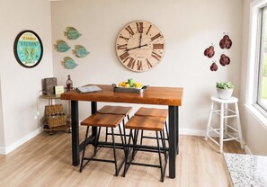 Uma área de refeições com uma mesa de madeira, banquetas de metal preto, um relógio de parede, arte de parede decorativa com peixes e um pequeno vaso com planta.