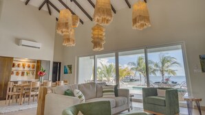 Een ruime woonkamer met een groot raam met uitzicht op een zwembad en palmbomen. Drie hangende rieten lampen, een houten eettafel met stoelen, en een groene bank.
