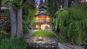 A two-story house with a stone fire pit in front, surrounded by a pond and dense greenery.