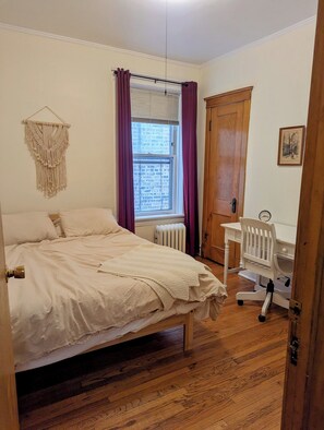 Une chambre à coucher comprenant un lit, un bureau, une chaise, un radiateur et une fenêtre avec des rideaux.