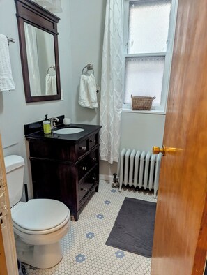 Une salle de bain comprenant des toilettes, un meuble-lavabo en bois foncé, un lavabo blanc, un miroir, des serviettes blanches, une fenêtre avec du verre dépoli, un radiateur et une porte.