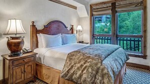 A bedroom with a large bed, wooden headboard, two bedside lamps, a nightstand with a telephone, and a window with a view of greenery.